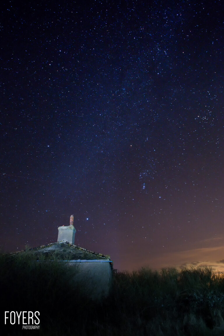 A lighthouse stands on a hill beneath a star-filled night sky, with grassland in the foreground and an orange glow on the horizon.