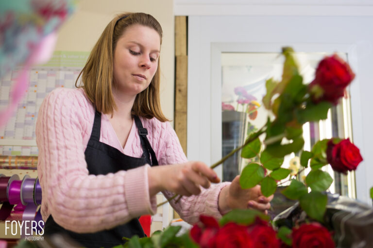 A florist in a pink cardigan and black apron arranges red roses at a work table in a flower shop.