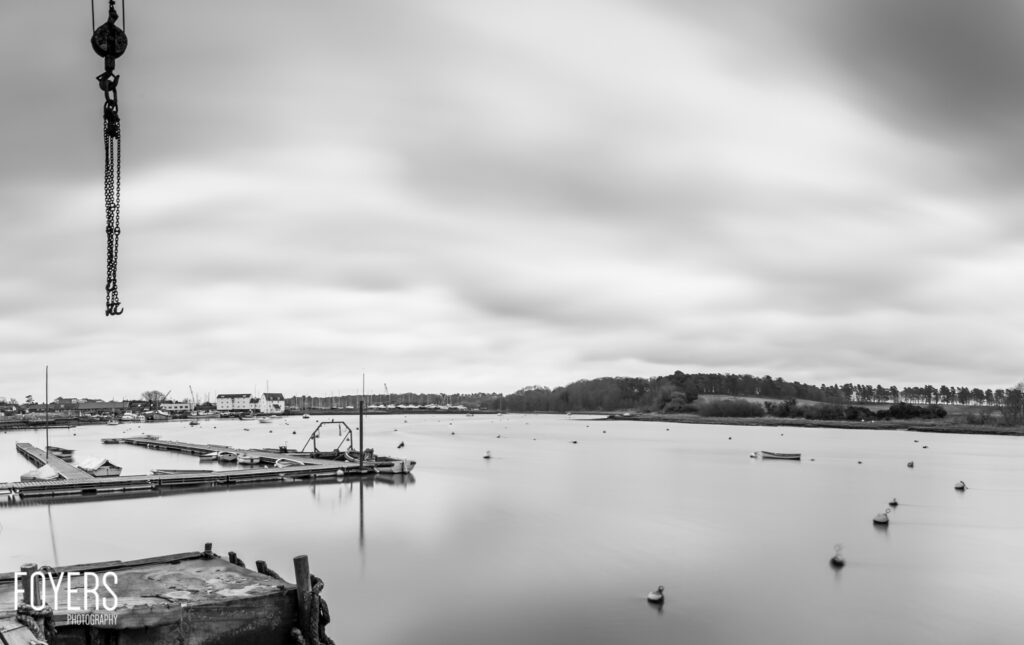 A chain and pulley hanging from above overlooks a moored sailing boat in a calm estuary under overcast skies.
