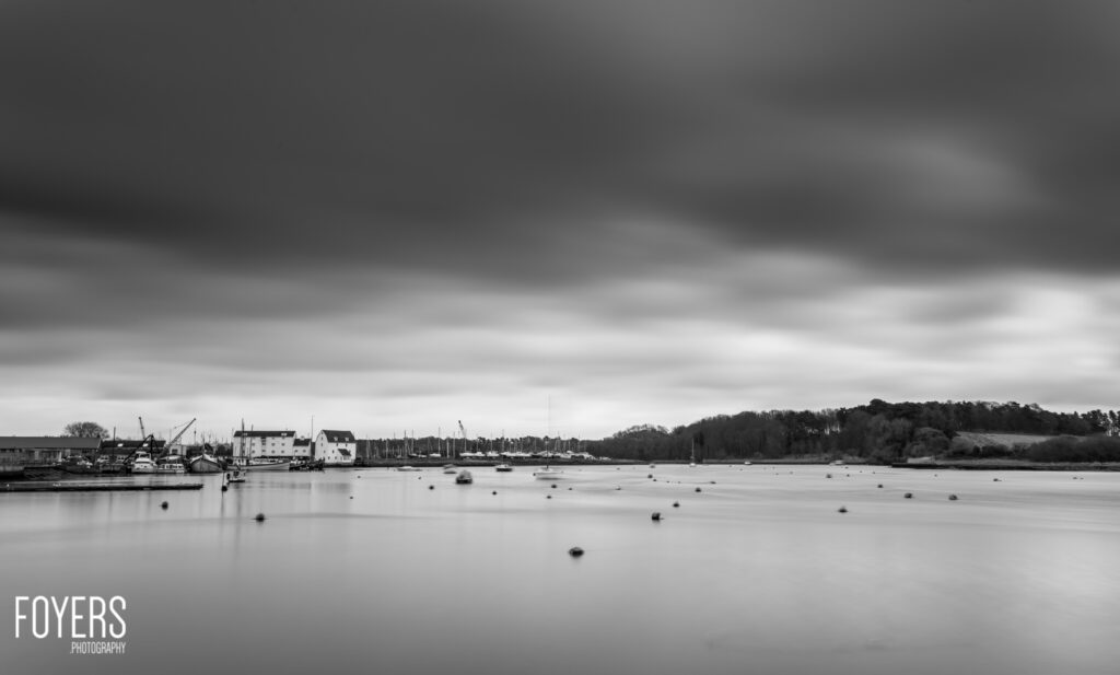 A mooring field with several small boats on calm water, waterfront buildings and trees visible across the harbour under overcast skies.