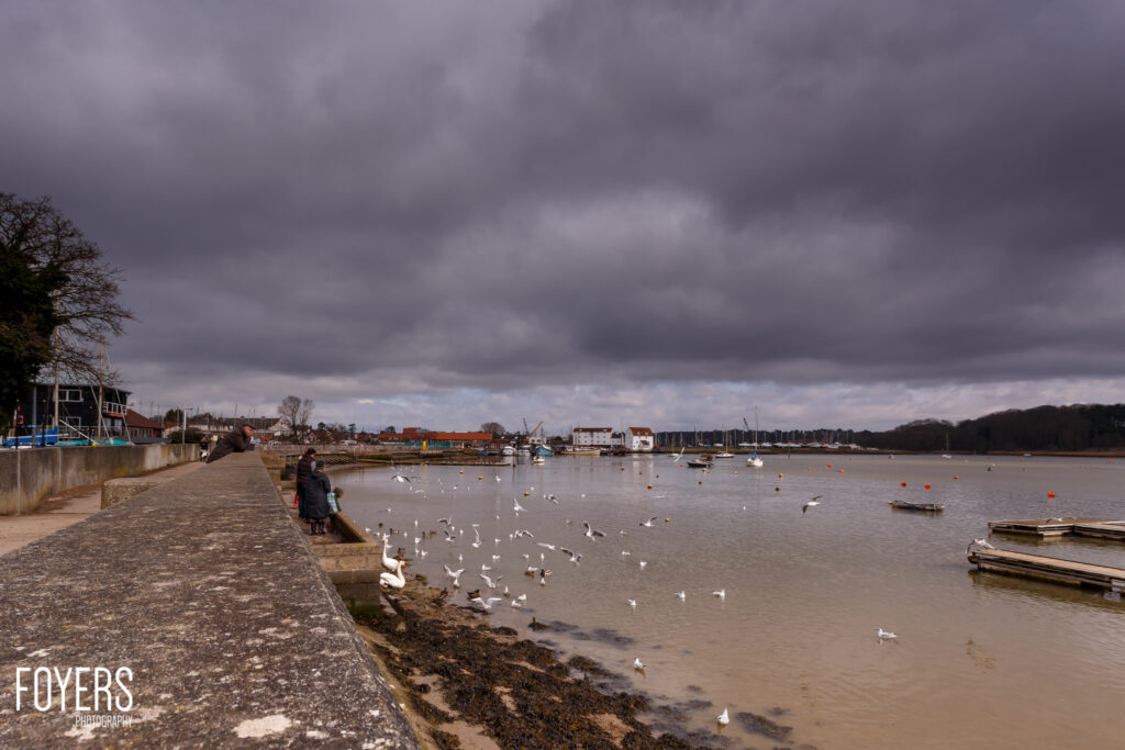 A waterfront promenade by a tidal river with moored boats and seagulls on the water under dramatic cloudy skies.