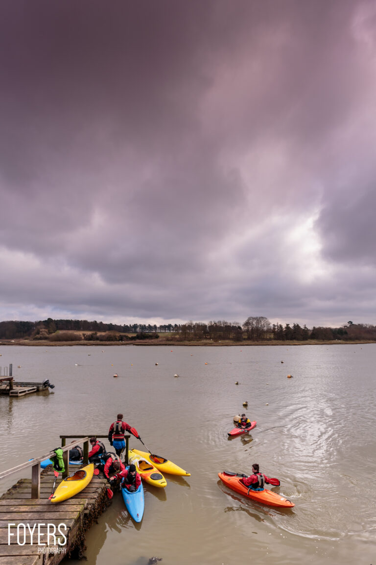 Kayakers in brightly coloured kayaks on a calm estuary. Some launch from a wooden jetty whilst others paddle in the shallow water.