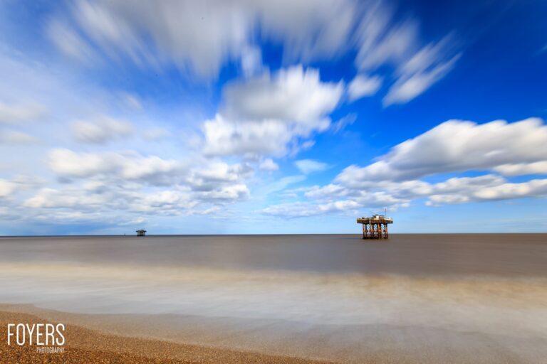 Beach scene with two abandoned wooden platforms in shallow water under a blue sky with white clouds.