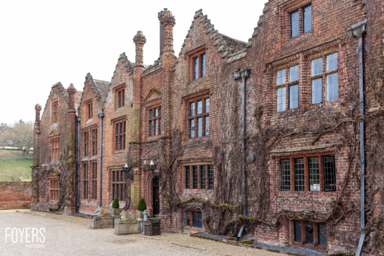A row of Victorian red brick buildings with distinctive chimneys and ivy-covered walls, set in a courtyard with gravel ground.