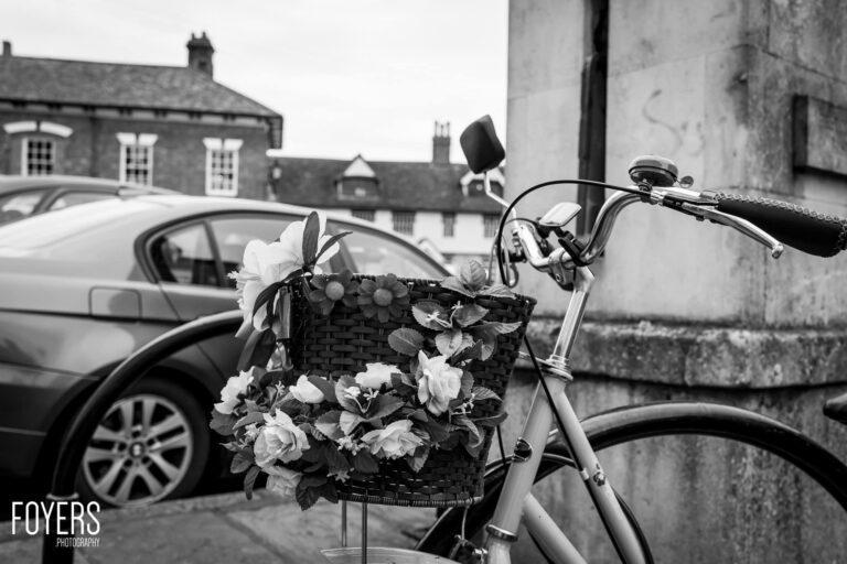 A vintage bicycle with a wicker basket overflowing with white flowers stands in front of a stone building and parked cars.