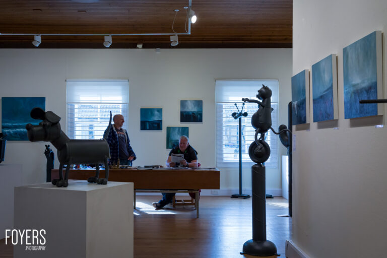 Art gallery interior with visitors viewing sculptures and blue seascape paintings on white walls under wooden ceiling.