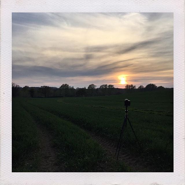 A tripod with camera stands in a green field at sunset, capturing the golden sun on the horizon beneath dramatic clouds.