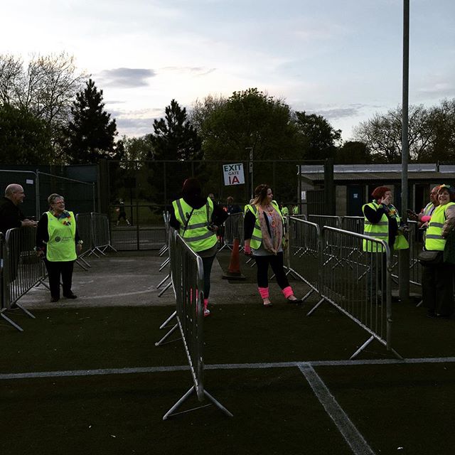 Runners in hi-vis jackets at a sports field event with barriers and officials during early evening.