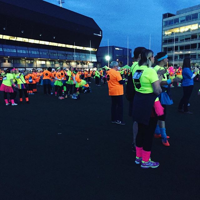 Large group of runners wearing bright neon orange and lime green tops gathered at dusk in an urban plaza before an event.