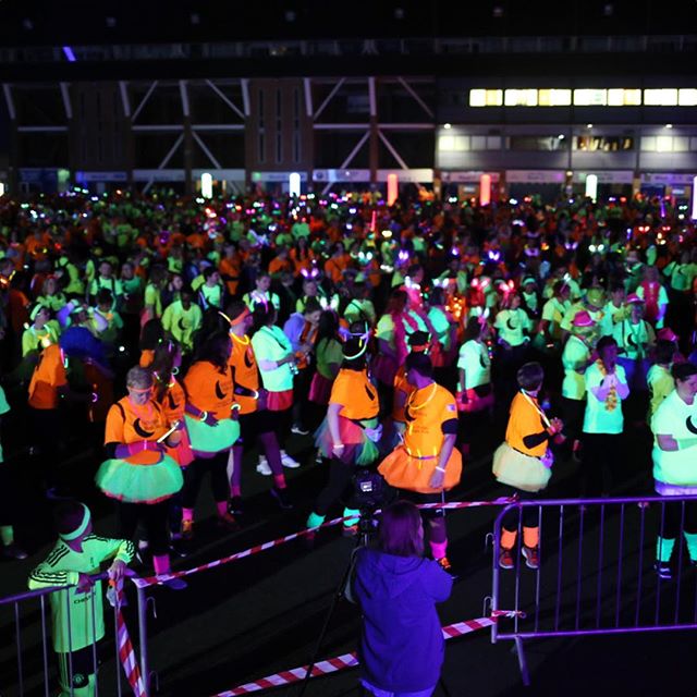 A large crowd of people wearing neon clothing and glowing accessories at a nighttime event in an indoor venue with industrial architecture.