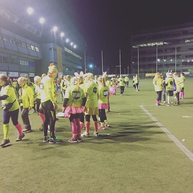 A large group of children wearing yellow and pink sports kits gathered on a floodlit football pitch at night, appearing to be at a youth sporting event.