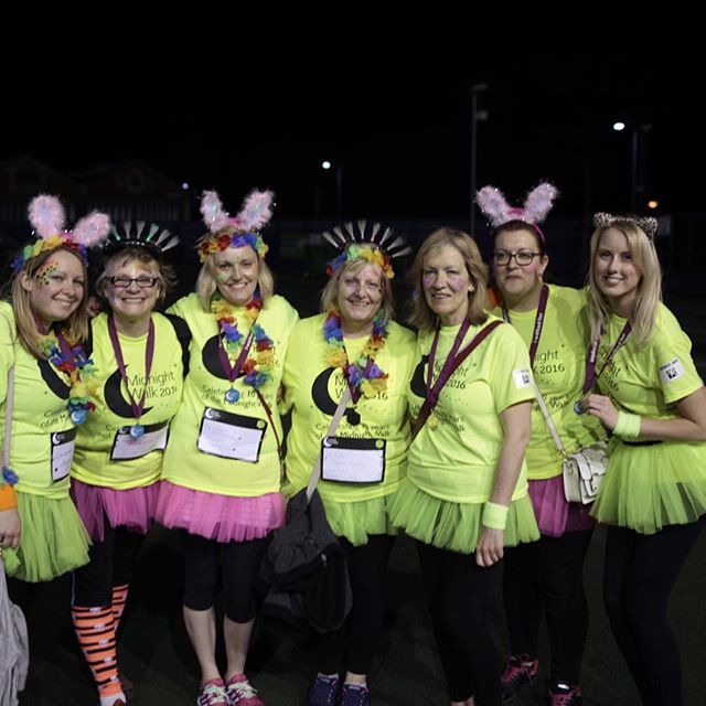Seven women wearing neon yellow shirts, tutu skirts and sparkly headbands pose together at night, smiling at the camera.