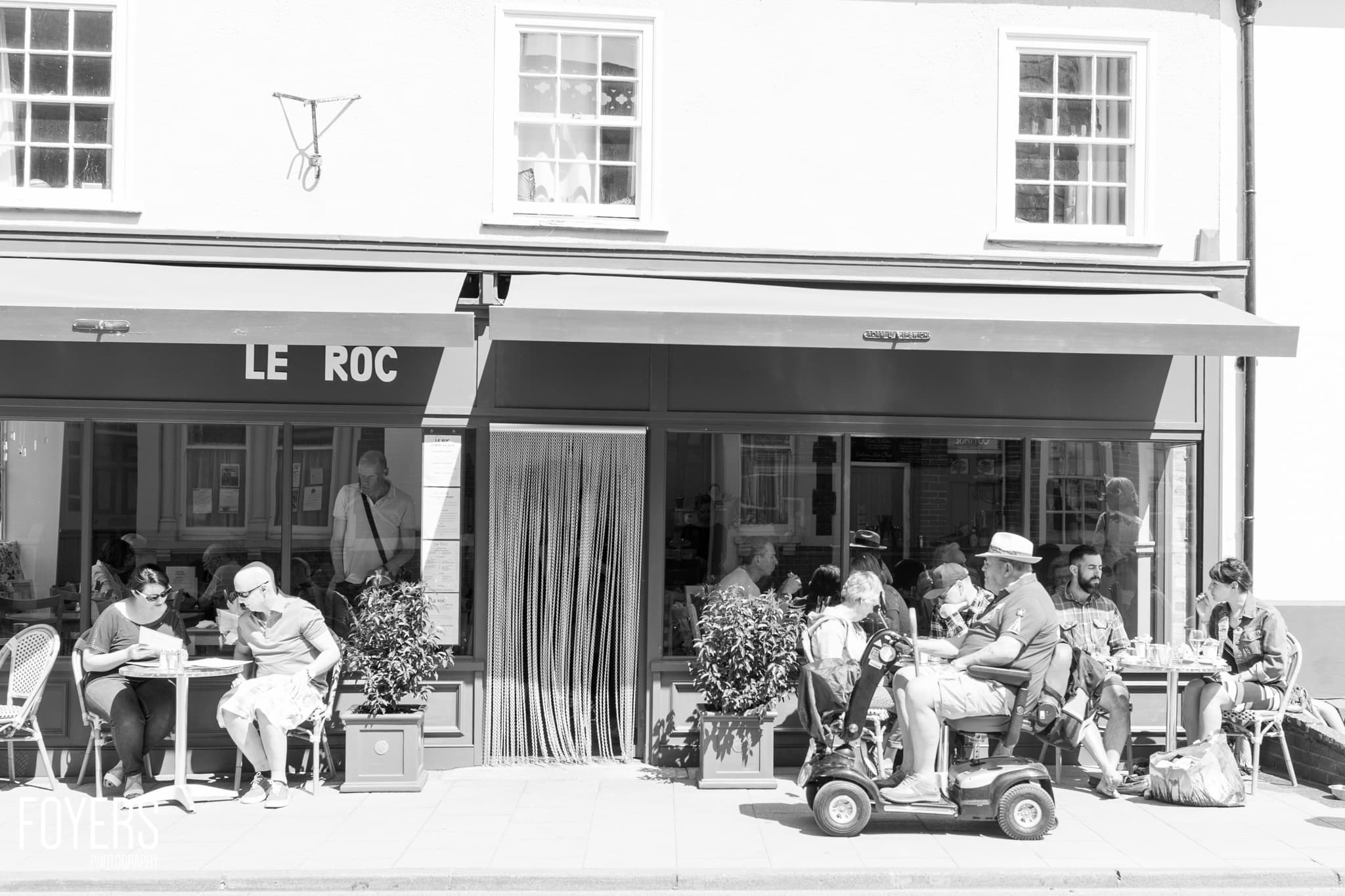 Black and white photo of a café called Le Roc with customers seated outside at tables on a sunny day.