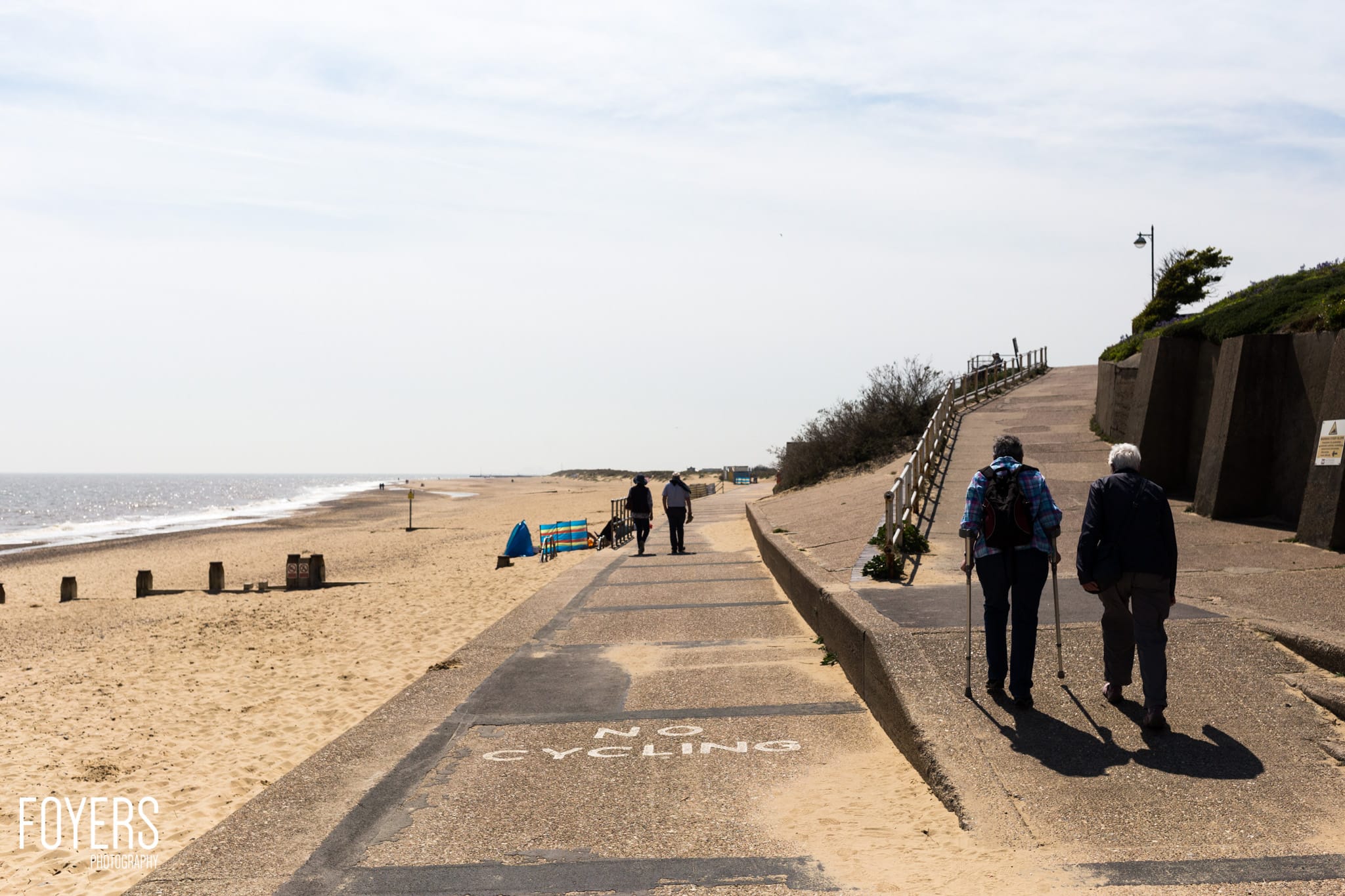 Two people walking on a promenade beside a sandy beach with sea defences and the ocean beyond on an overcast day.