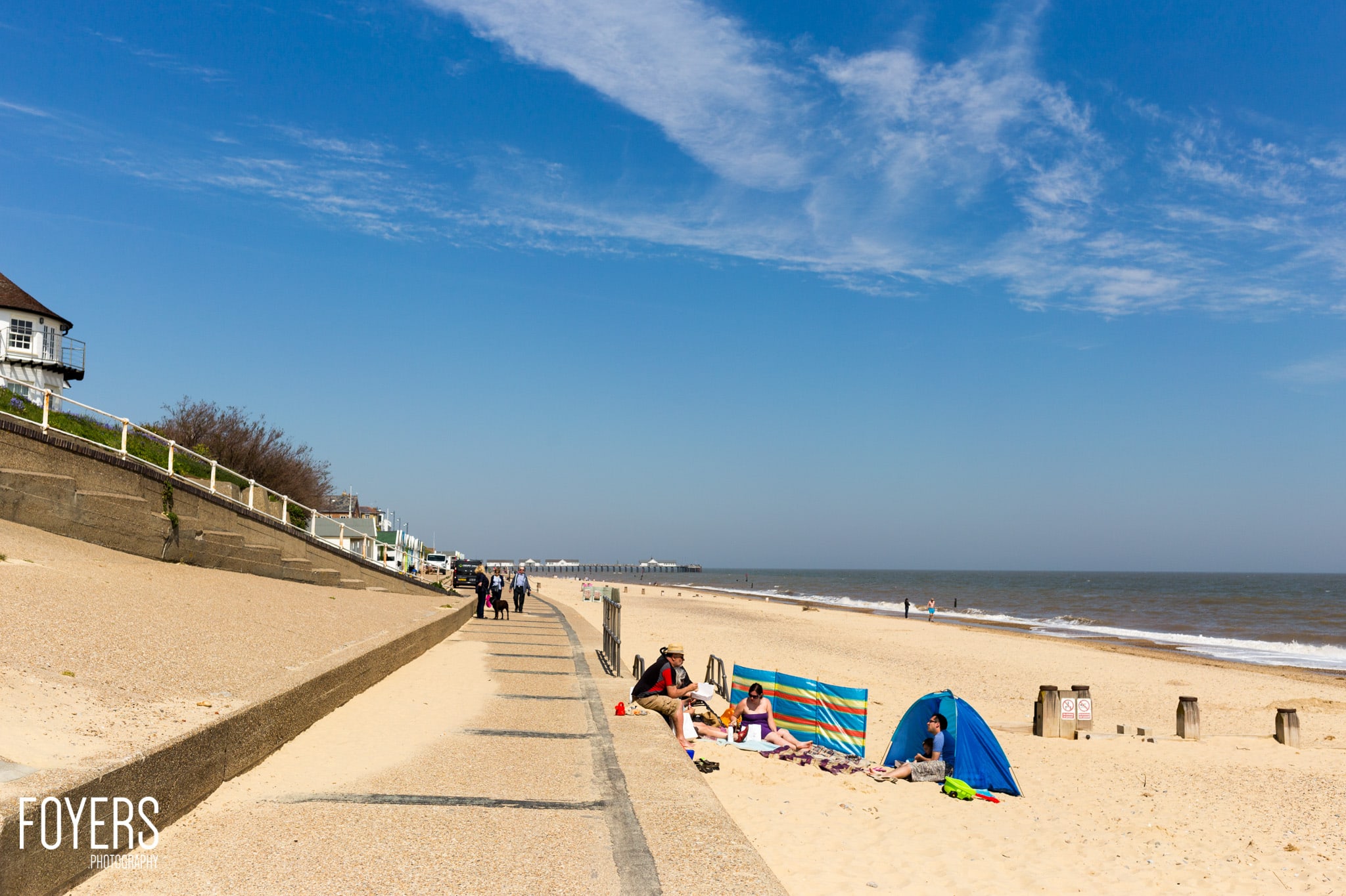 Beach scene with beachgoers, colourful windbreak, blue tent, wooden sea defences and pier visible in distance under clear sky.