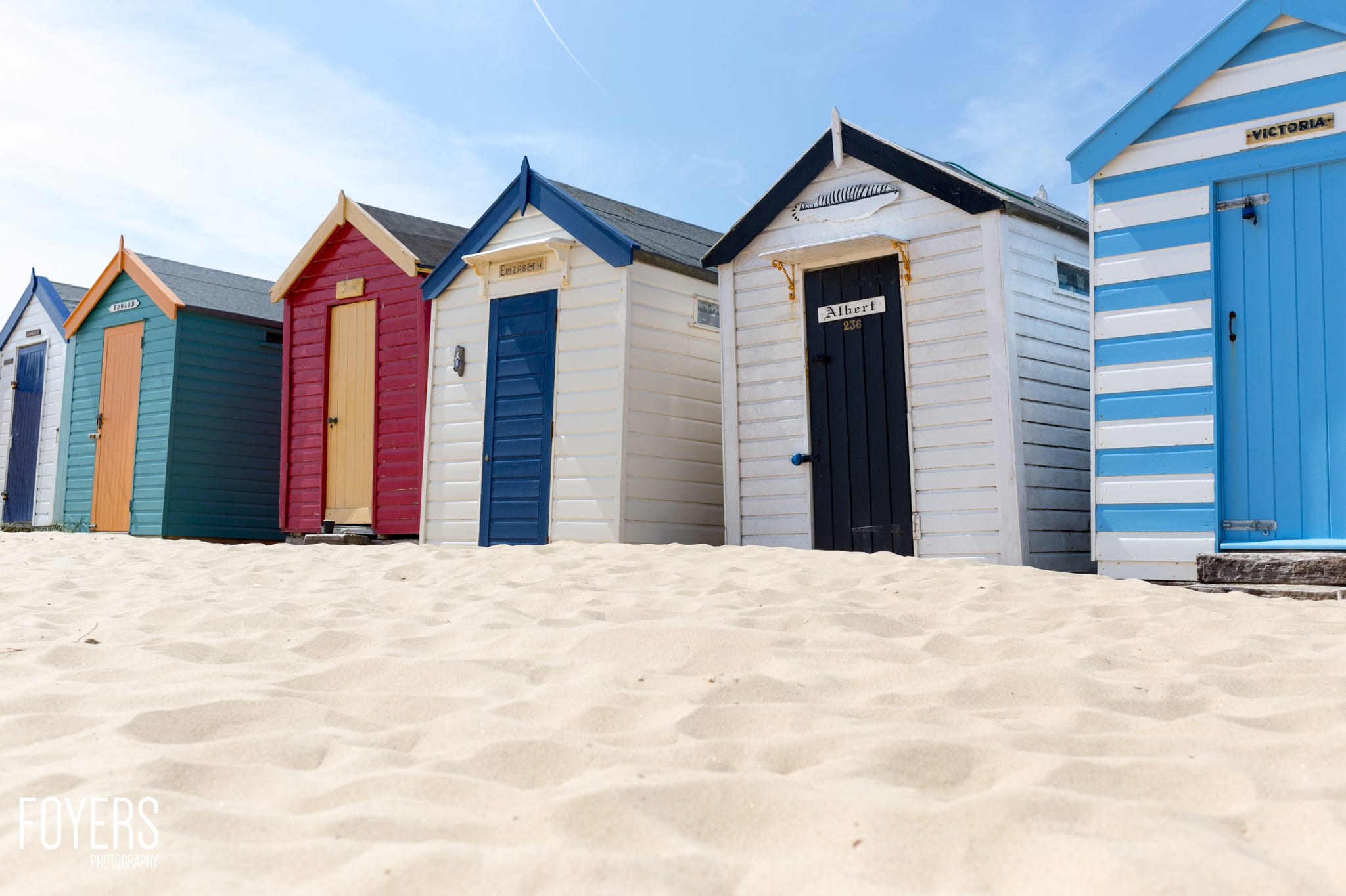 Colourful beach huts lined up on sand under a blue sky. Each hut has a different colour scheme and pitched roof design.