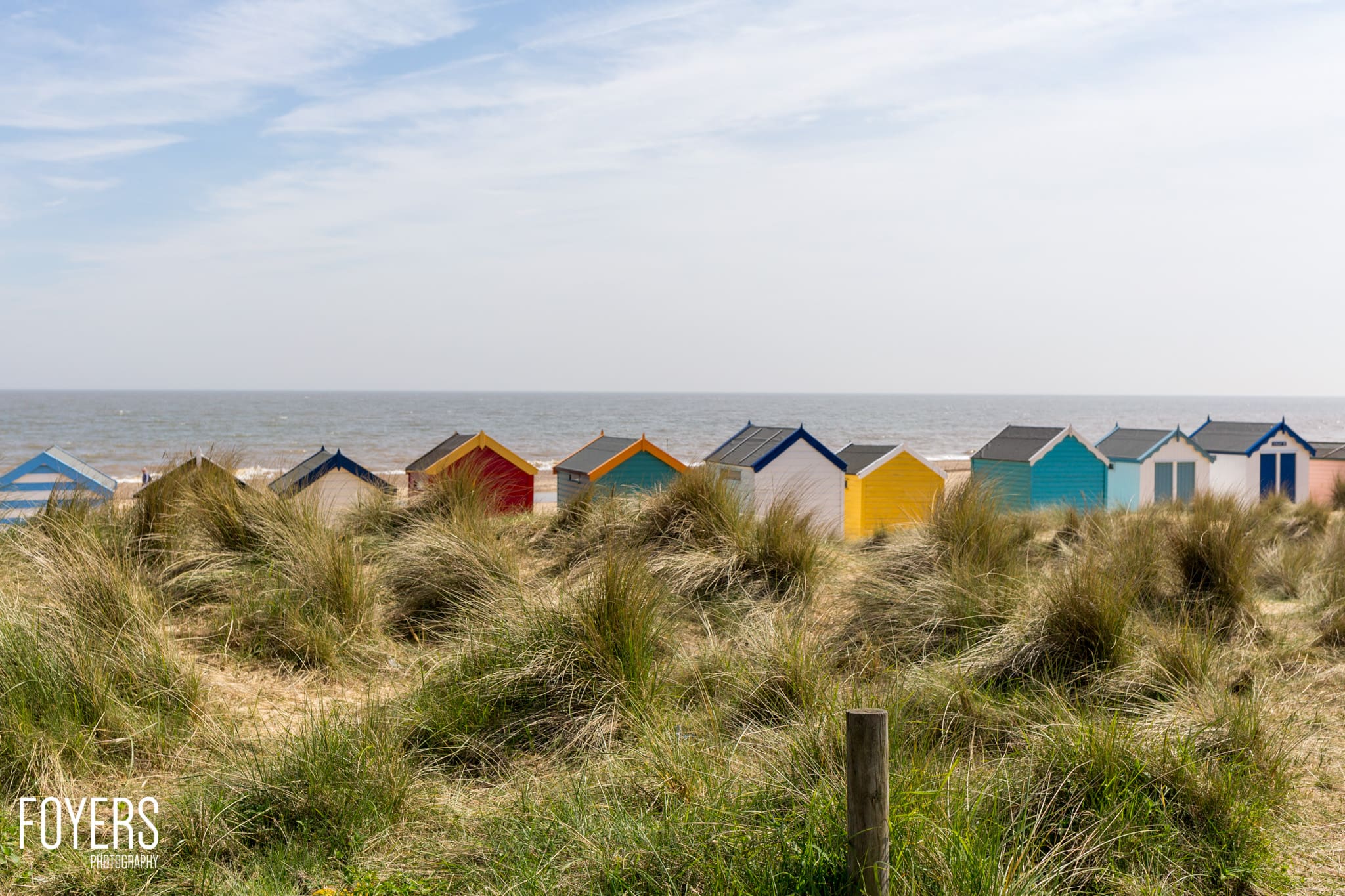 Colourful beach huts line a sandy shore backed by dunes with marram grass, sea and sky beyond.