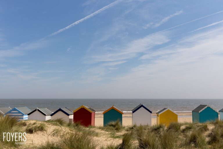 Colourful beach huts lined up on sandy dunes facing the sea under a blue sky with wispy clouds.