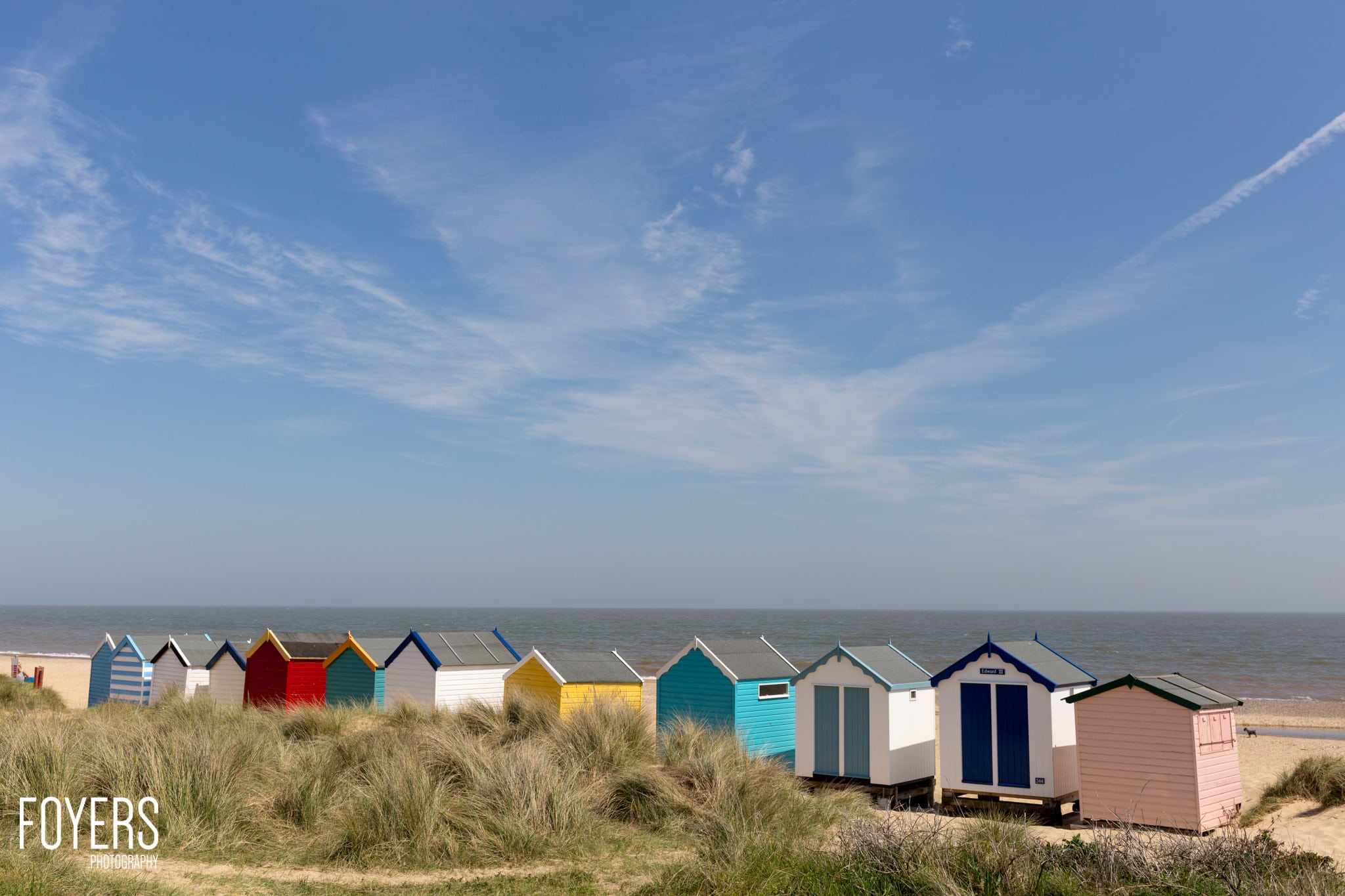 A row of colourful beach huts lined up on sandy beach with marram grass, sea and sky behind.