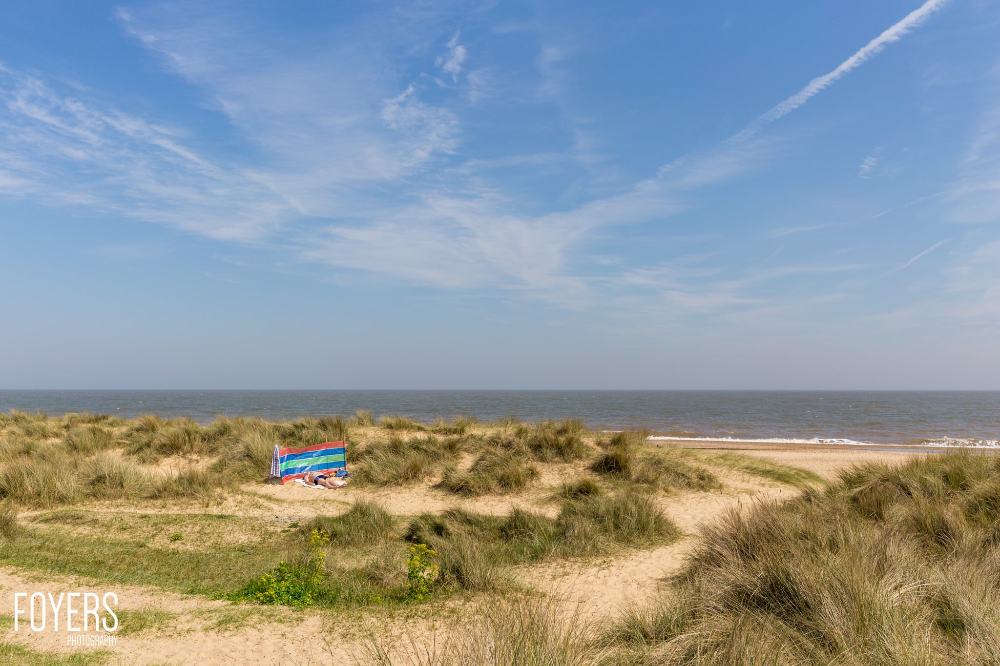 A person lies on a colorful beach towel in the sand dunes overlooking a calm sea under a blue sky with wispy clouds.