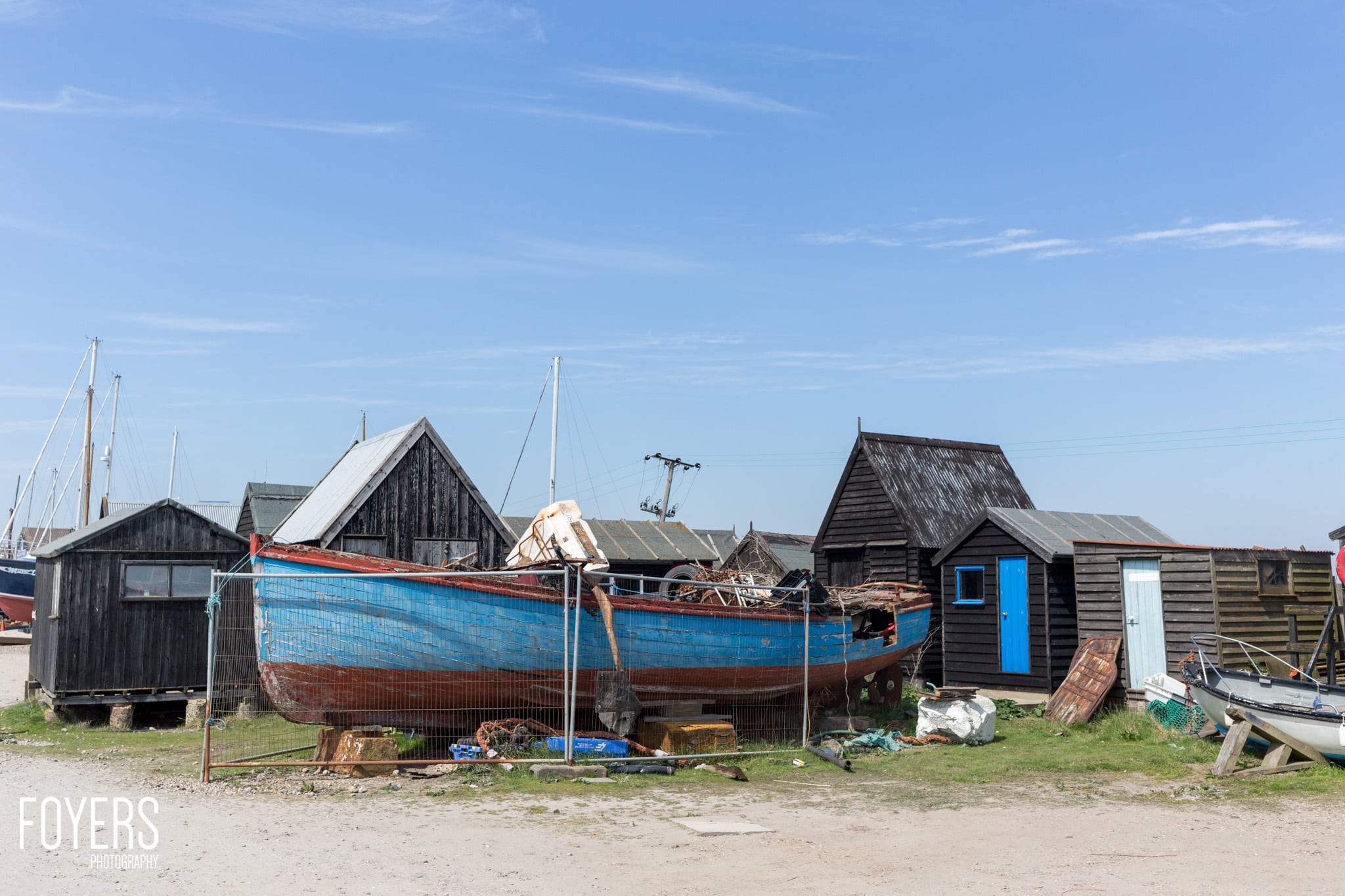 A beached fishing boat with blue and red hull surrounded by weathered wooden boathouses on shingle beach under clear sky.