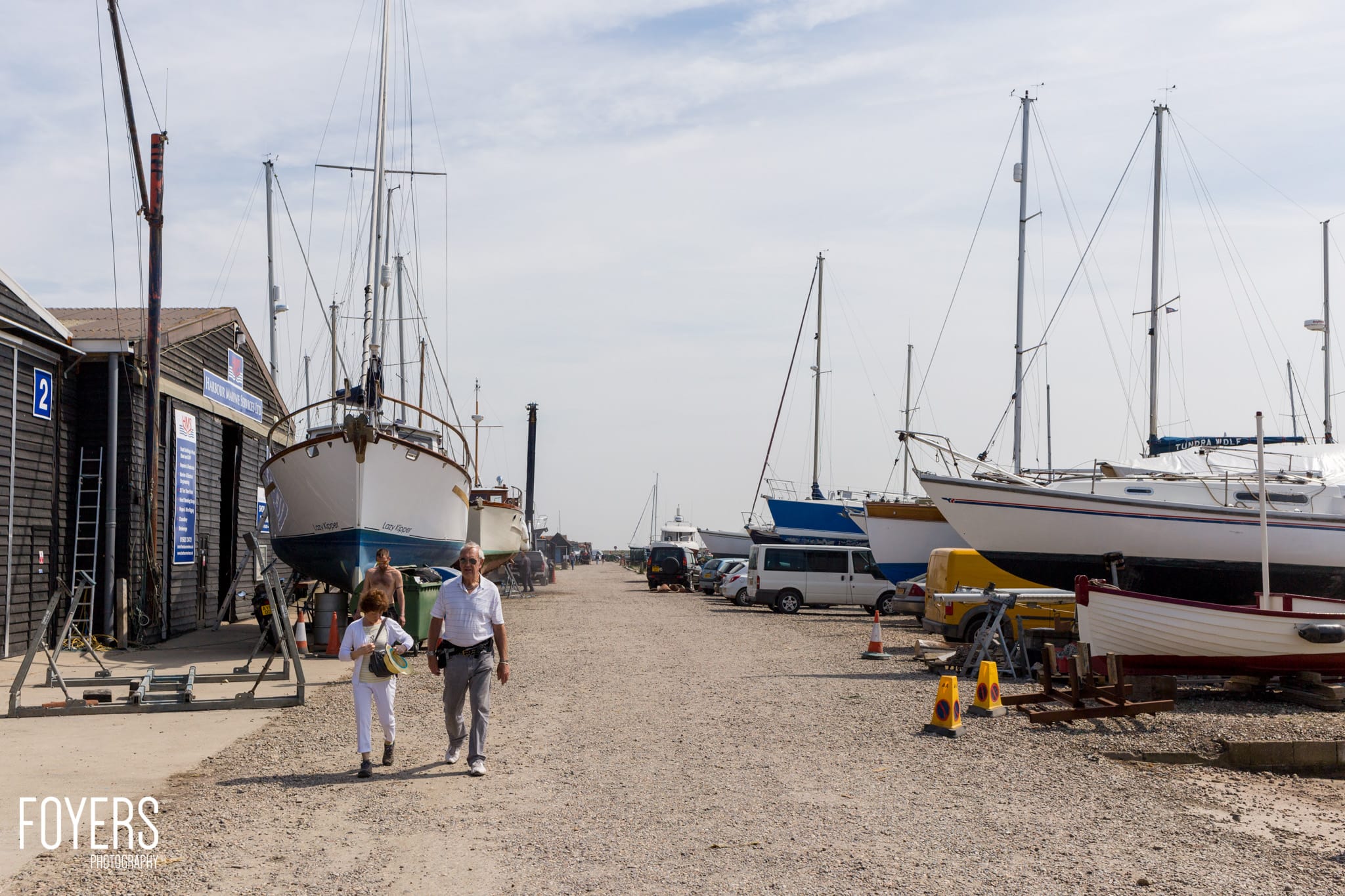 A man and child walk along a sandy beach lined with moored sailing boats and wooden sheds on a cloudy day.
