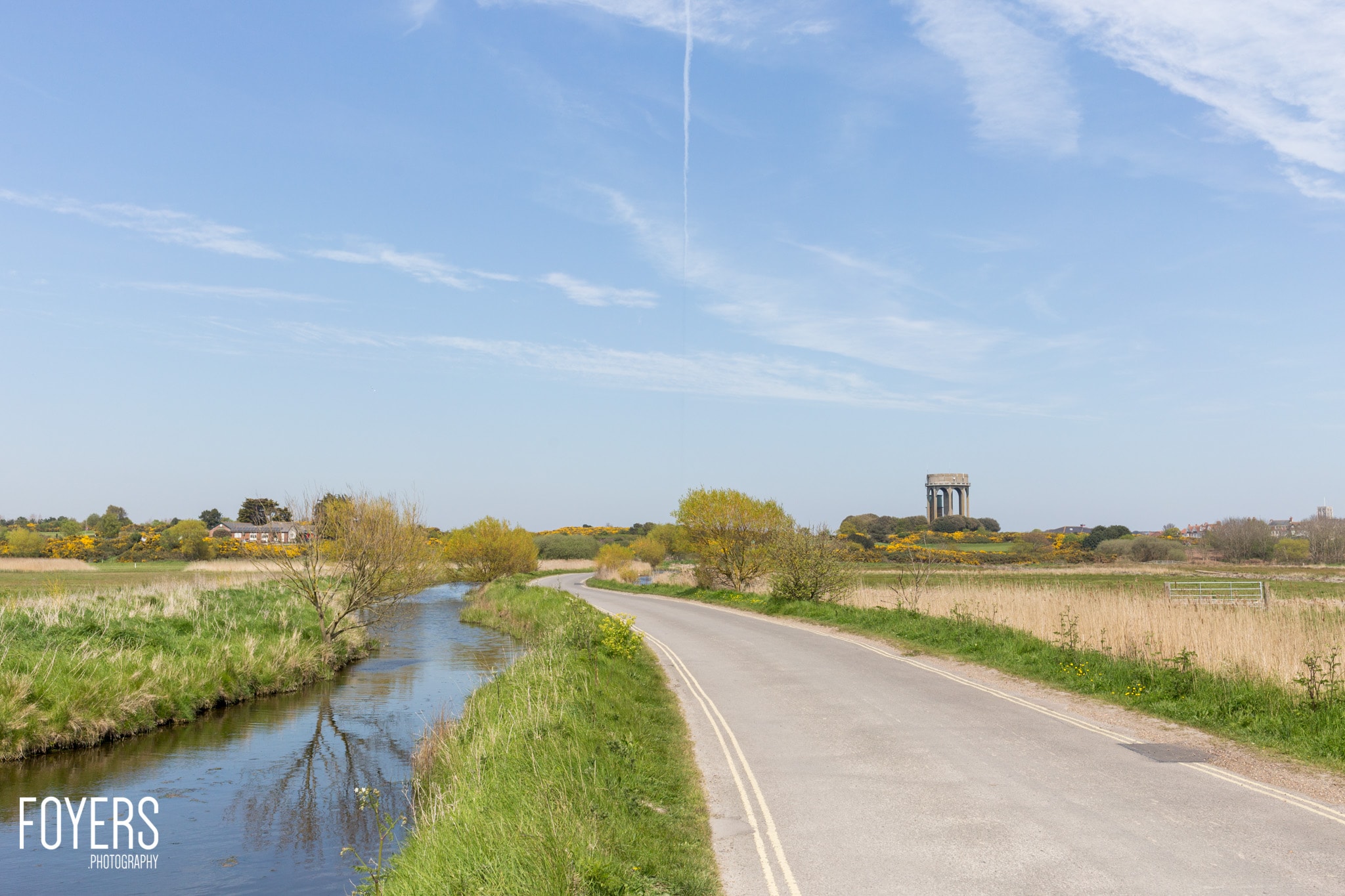 A straight rural road runs alongside a drainage ditch under a blue sky, with fields and trees stretching to the horizon.