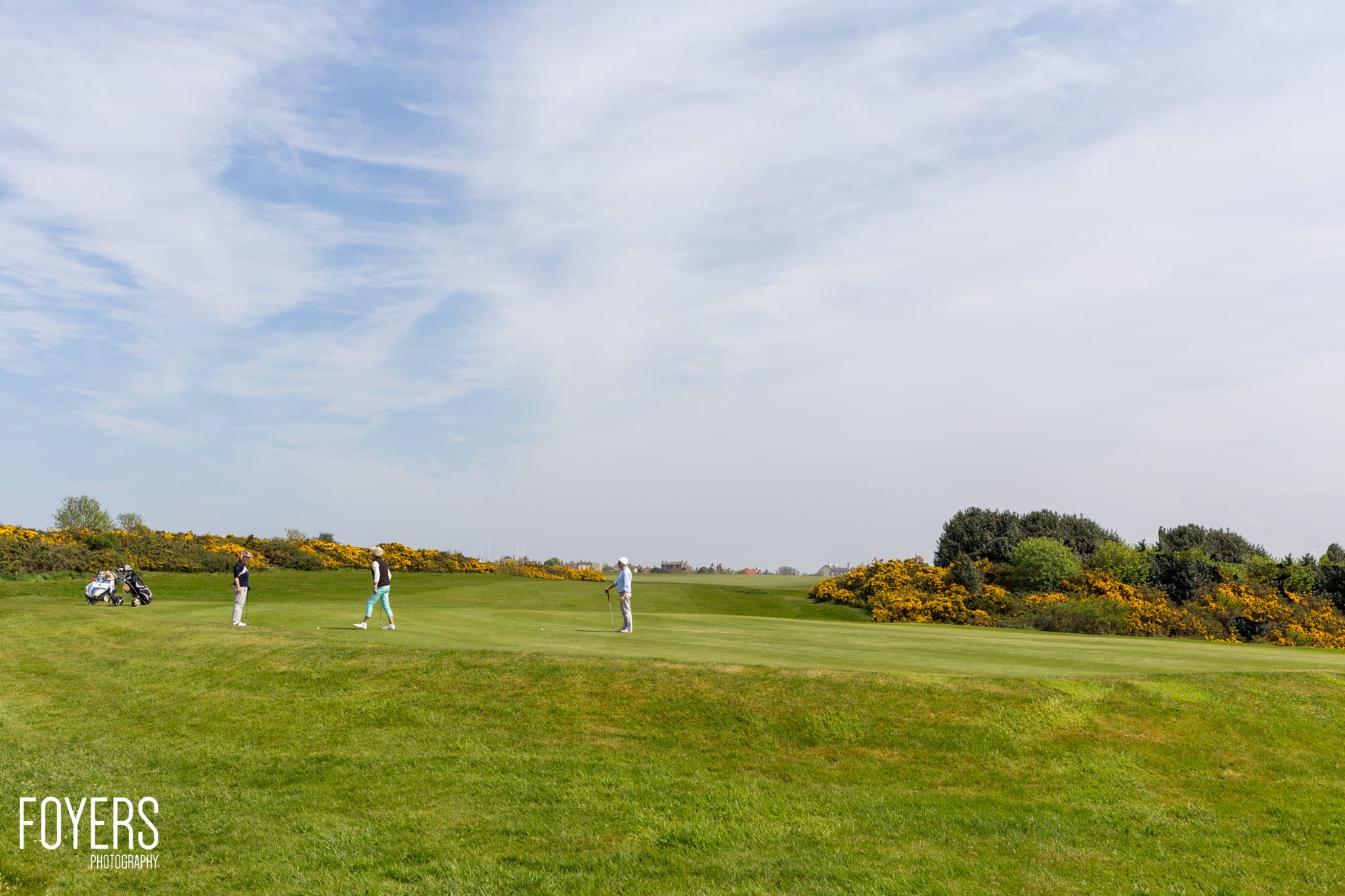 Golfers playing on a large green fairway with yellow gorse bushes lining the course under a partly cloudy sky.