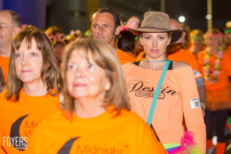 A woman in a cowboy hat and peach-coloured top stands among a crowd wearing orange shirts at a nighttime outdoor event.