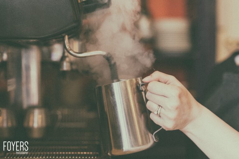 A person holding a metal milk jug with steam rising, wearing a ring, in what appears to be a café setting.