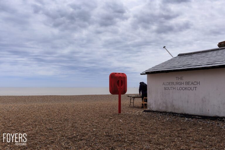A person sits outside the Aldeburgh Beach South Lookout hut on a pebble beach under a cloudy sky, next to a red telephone box.