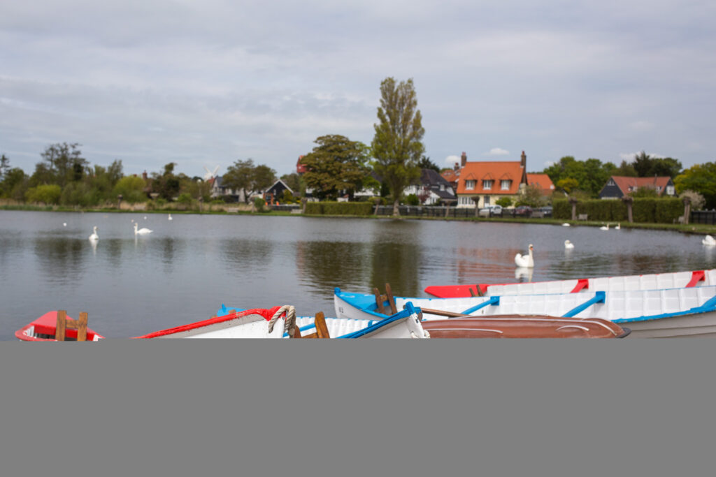 Rowing boats moored on Thorpeness Meare, with swans on the water and a house among trees behind