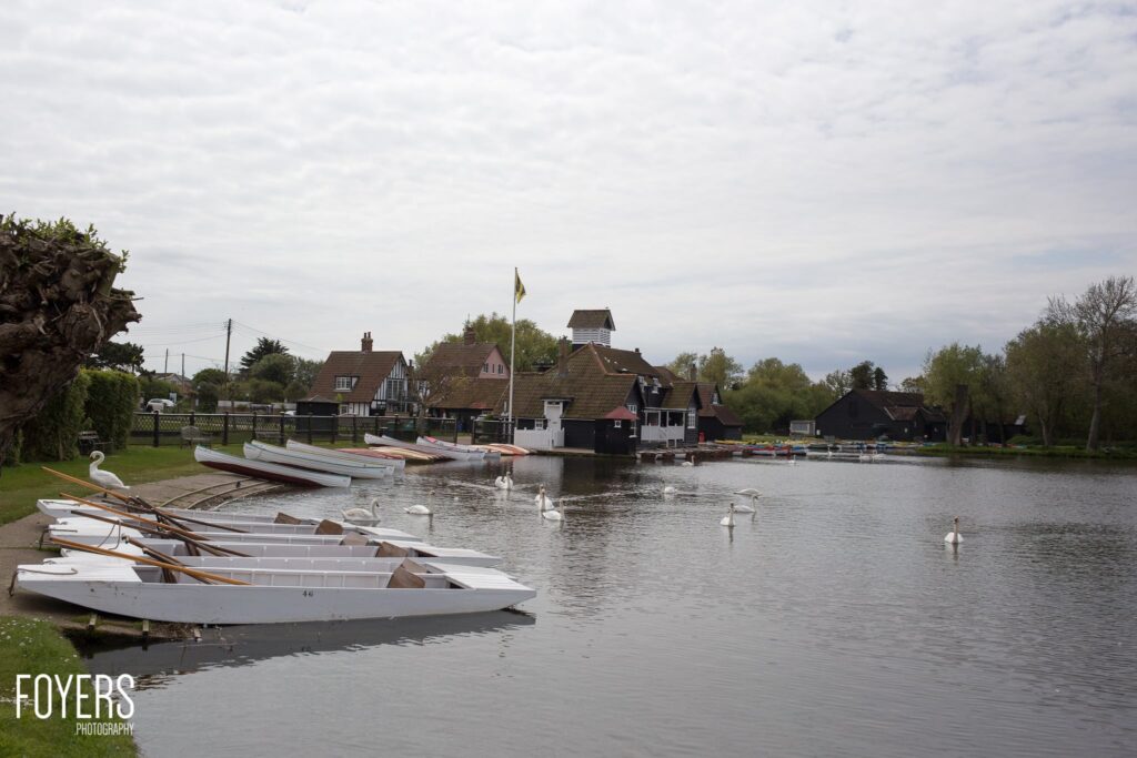 Rowing boats moored on a riverbank beside a grassy verge, with traditional buildings and swans on the water beyond.