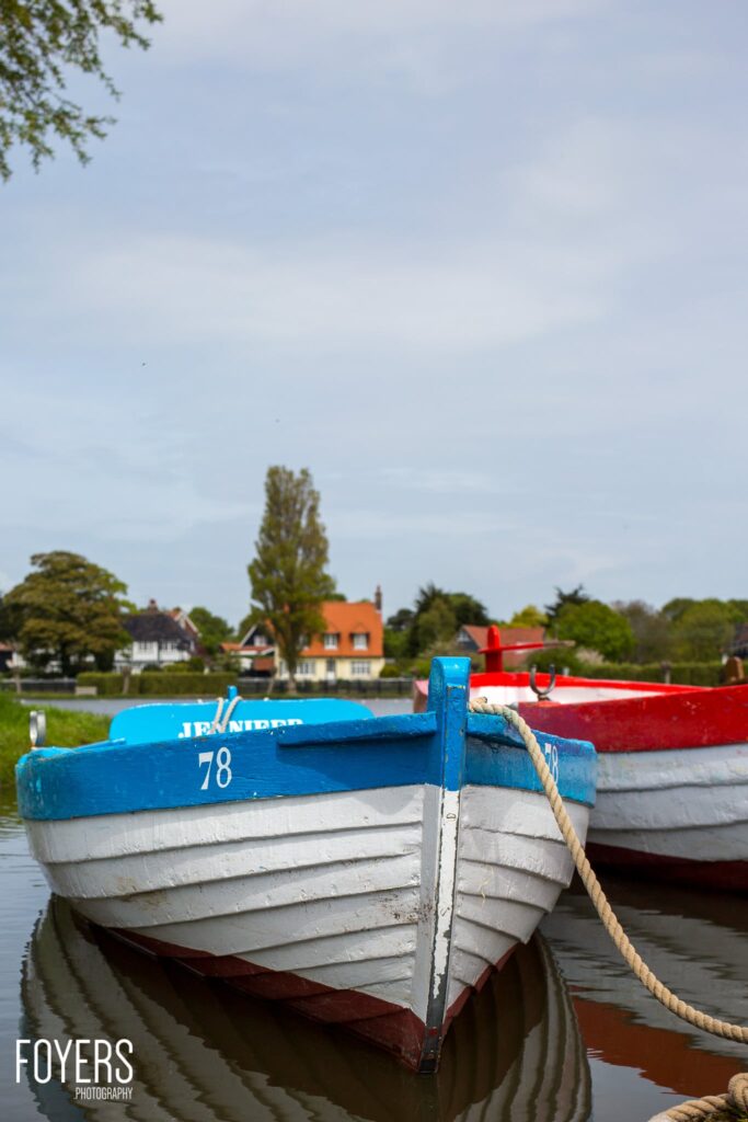 Moored rowing boat numbered 78 with blue and white hull on calm water, with cottages and trees in background.