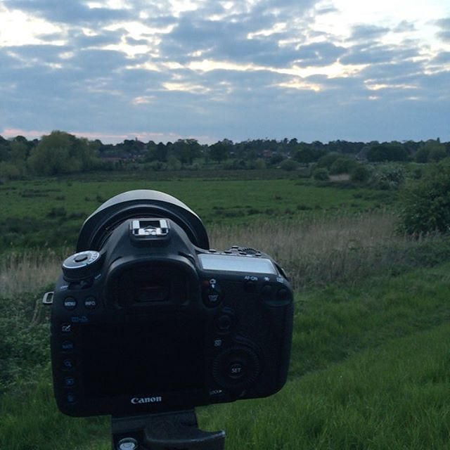 A Canon DSLR camera on a tripod pointed at a rural landscape with green fields, trees, and a cloudy sky.