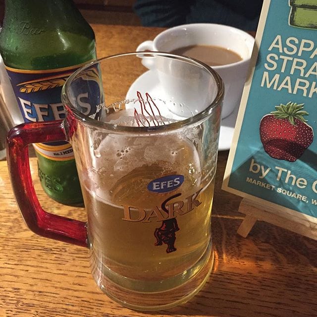 A glass mug of Efes Dark beer sits on a wooden table next to a green bottle and a white cup of coffee.