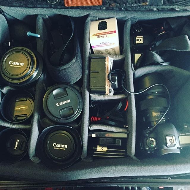Camera equipment organised in a padded case, including Canon lenses, bodies, batteries, chargers, and accessories neatly arranged in compartments.