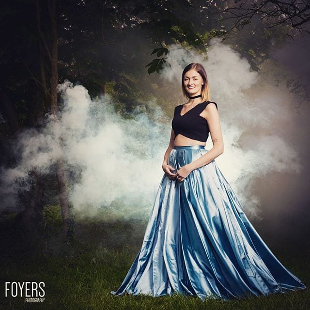 A woman in a black crop top and flowing blue skirt stands smiling in a misty forest setting with dramatic white smoke.