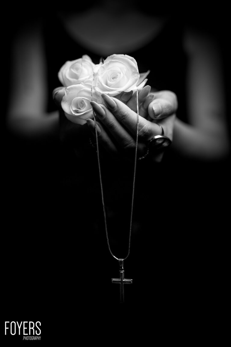 Hands holding white roses with a cross pendant necklace against a dark background. Black and white photograph.