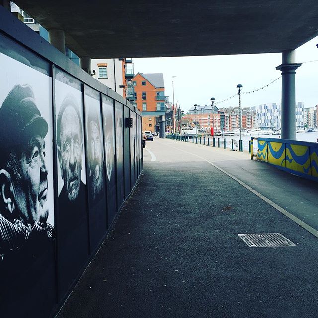 A pedestrian underpass with black and white portrait panels on the left wall, leading toward a waterfront with moored boats and brick buildings.