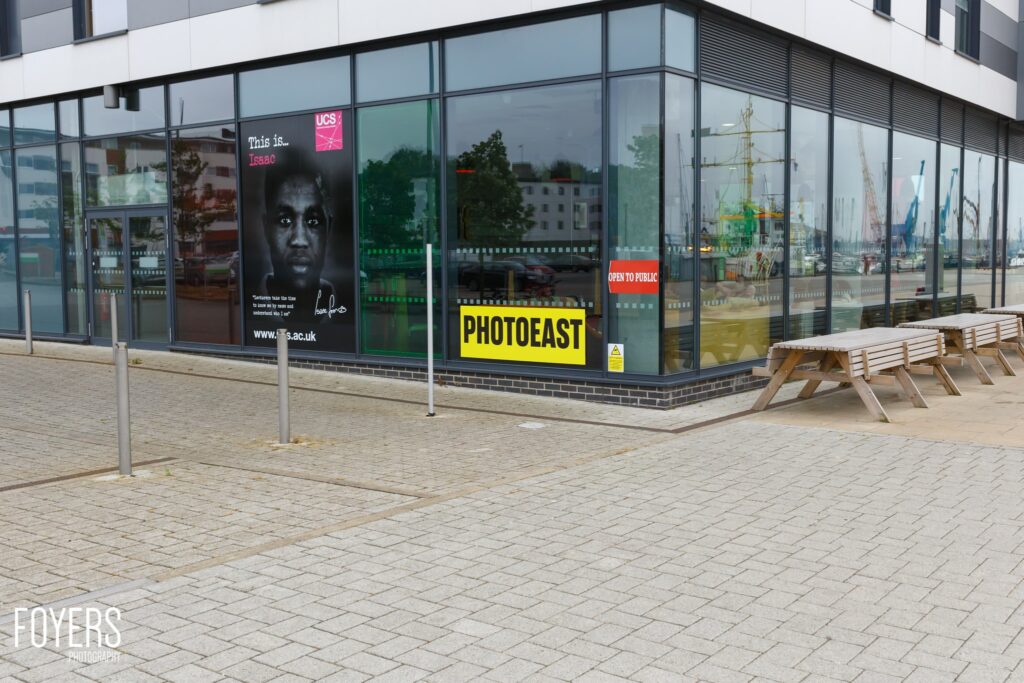 A modern glass-fronted gallery space displays exhibition posters, including one reading PHOTOEAST in yellow text. The space is open to the public.