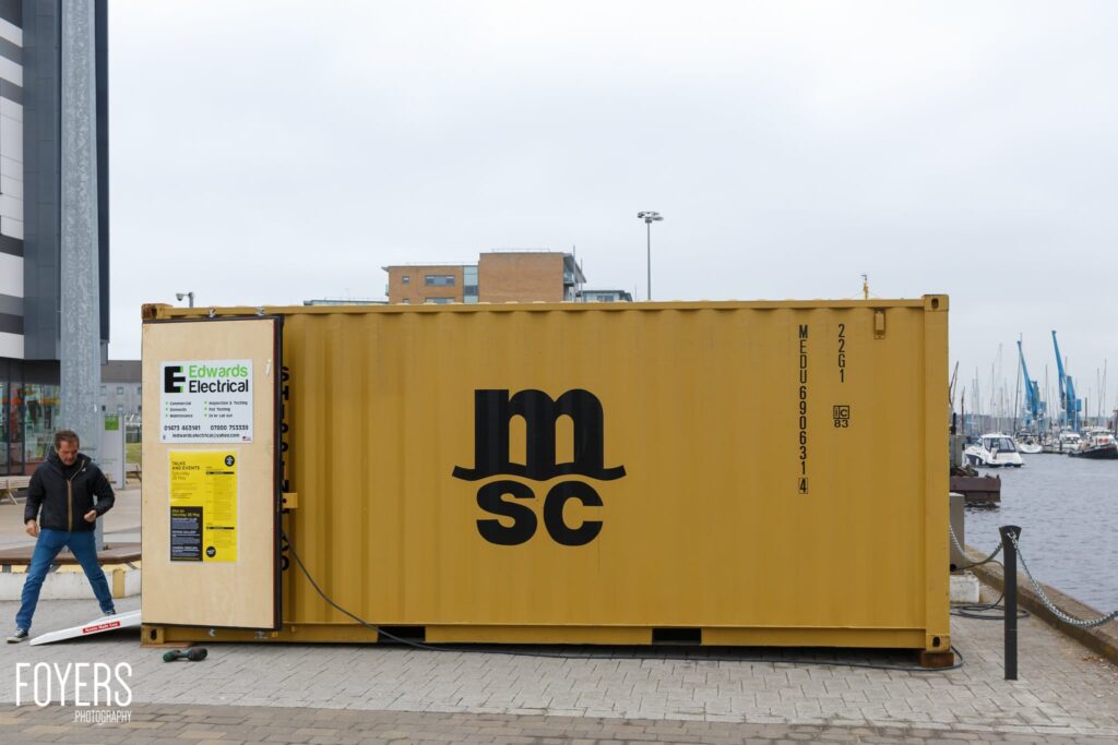 A yellow shipping container with msc logo sits on a waterfront promenade next to buildings and moored boats.