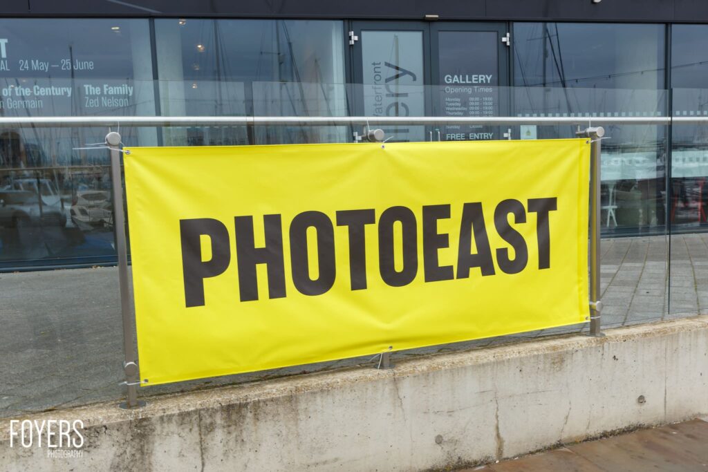 A bright yellow banner reading PHOTOEAST hangs on a gallery storefront with glass windows and opening times displayed.