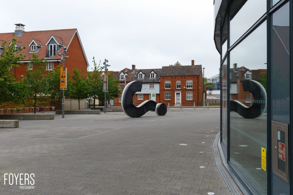 A modern black sculptural installation in a town square, surrounded by historic brick buildings and paved plaza.