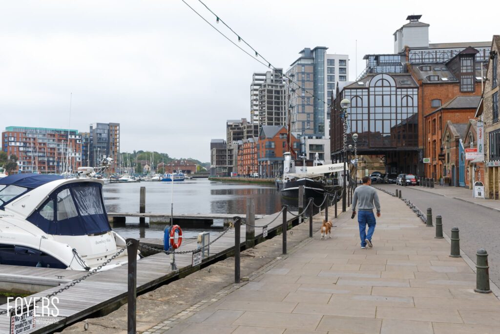 A man jogs along a waterfront promenade with his dog, passing moored boats and Victorian brick buildings in a coastal town.