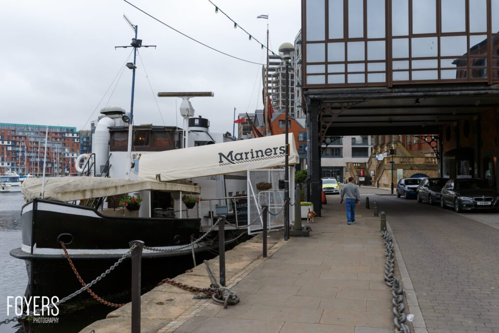 A moored fishing vessel named Mariners is docked at a waterfront promenade with modern buildings and shops visible behind it.