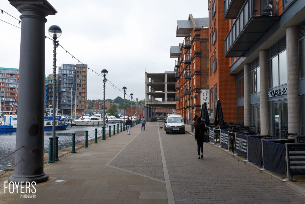 A waterfront promenade with brick buildings, lamp posts, and a few pedestrians. Boats visible on the left by the water.