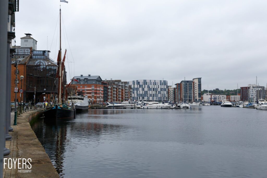 A waterfront harbor with moored boats, historic and modern buildings reflected in calm water under overcast sky.
