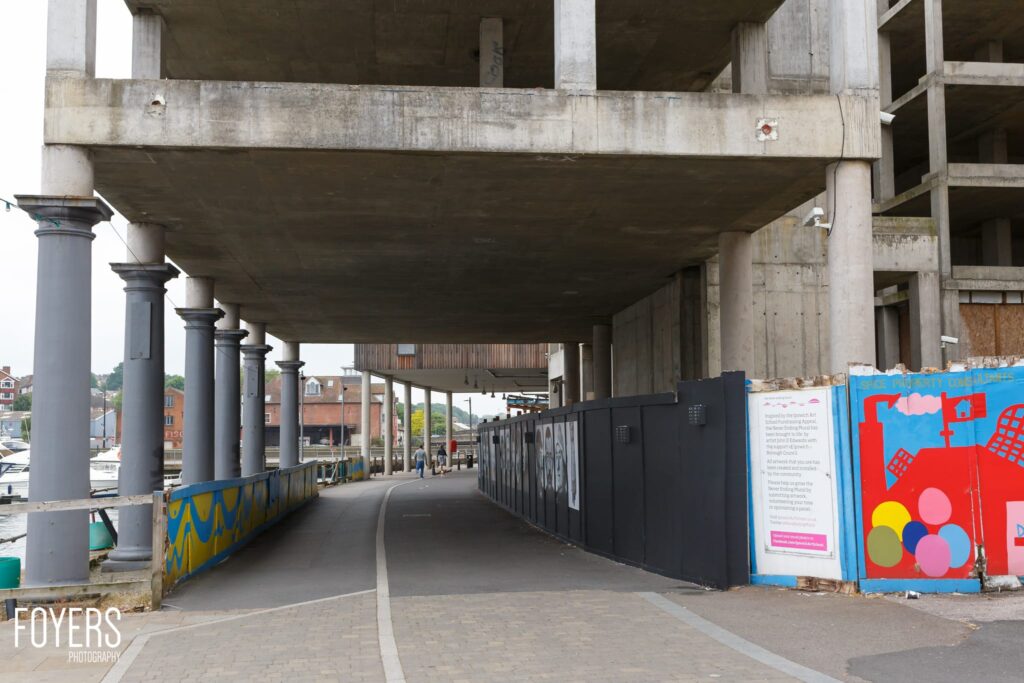 Underpass with concrete pillars, colorful street art panels on right, brick buildings visible beyond.