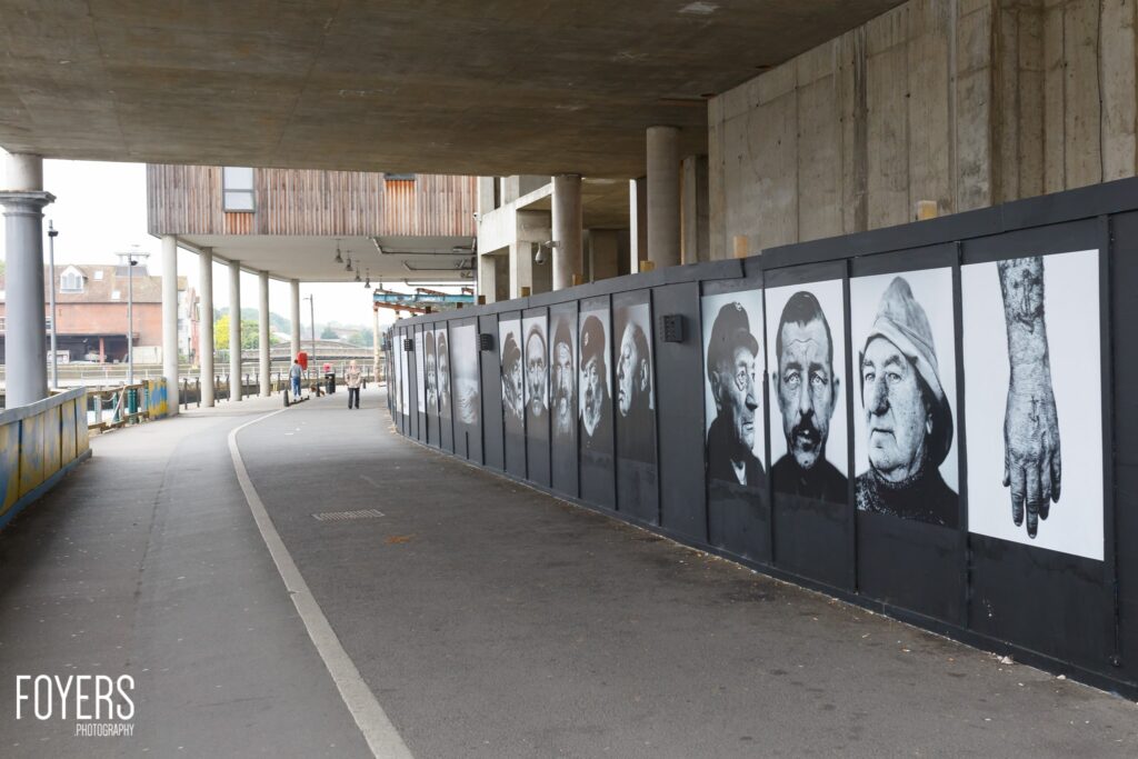 A pedestrian underpass beneath a concrete bridge displays a series of black and white portrait photographs mounted on a dark panel along the right wall.